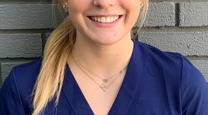 blond, green-eyed woman with hair pulled in ponytail and headband standing in front of grey brick wall and wearing blue "Southern Eye Bank" scrubs